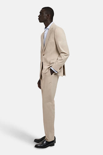 Pantalon de costume beige MANGO, dans une couleur unie, vu de côté avec une veste coordonnée et des chaussures noires.