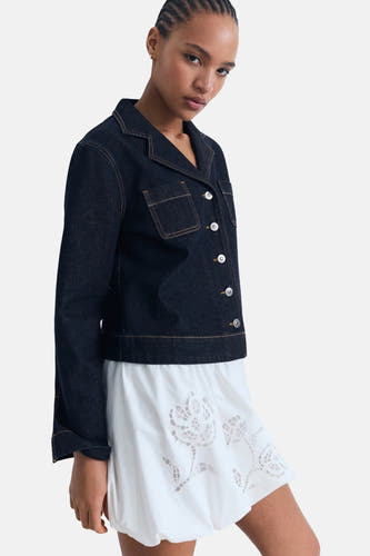 Veste en jeans bleu marine de MANGO, avec des manches longues et des boutons blancs, associée à une robe blanche brodée.