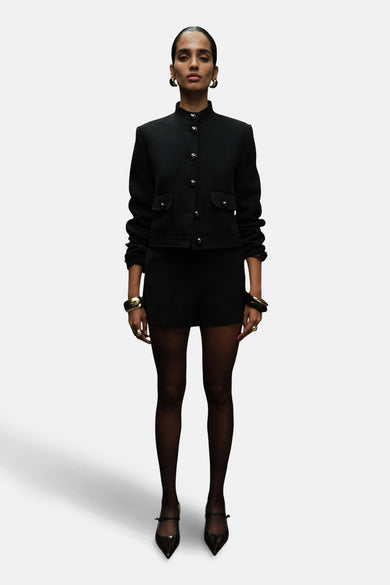 Blazer noir de MANGO, avec des boutons dorés et des poches brodées, porté avec des short noirs et des slips.