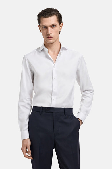 Chemise blanche à manches longues de MANGO, portée avec un pantalon bleu marine et une main dans la poche.