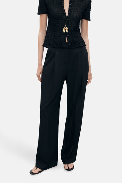 Pantalon noir de MANGO, avec plis et une large ceche, associé à un blouson noir et une chaîne dorée.