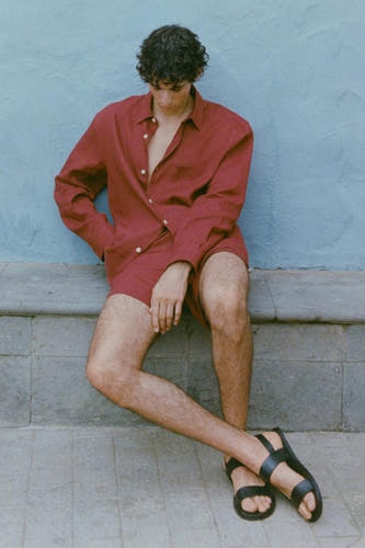 Chemise rouge à manches longues de MANGO, portée avec des shorts et des sandales, assise contre un mur bleu.