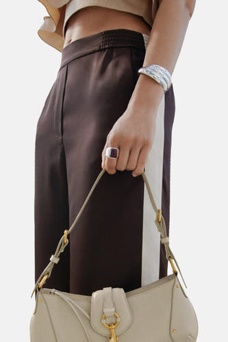Pantalon marron de MANGO, avec une bande blanche sur le côté, associé à un sac beige et un bracelet argenté.