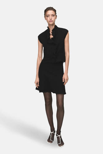 Jupe courte noire de MANGO, avec une finition texturée et un col en biais froncé, portée avec des collants noirs et des sandales blanches.
