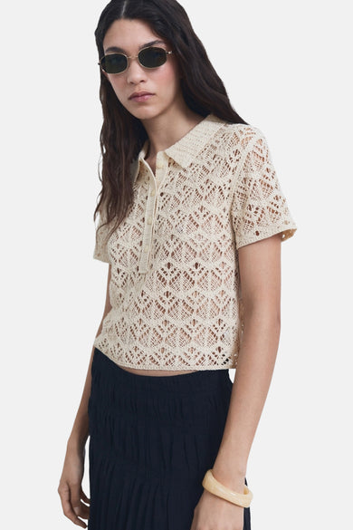 Gilet beige à col rond de MANGO, avec un ajour ouvert, porté avec une jupe sombre et un bracelet voyant.