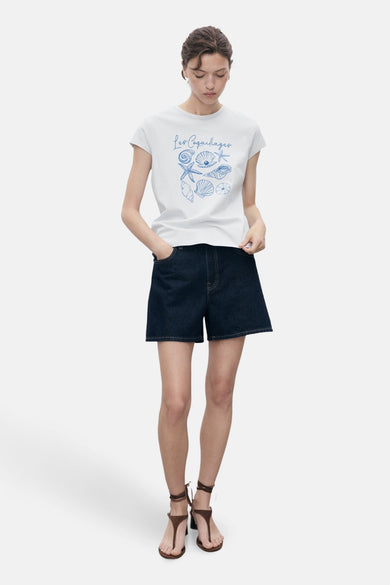 T-shirt bleu à manches courtes de MANGO, avec une impression de coquillages bleus et porté avec des shorts en denim et des sandales.