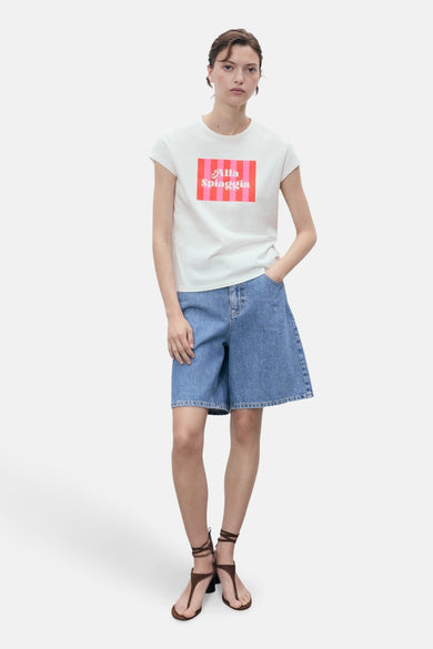 T-shirt blanc à manches courtes de MANGO, avec du texte 'Alla Spiaggia' rose sur la poitrine, associé à des bermudas en denim et des sandales.