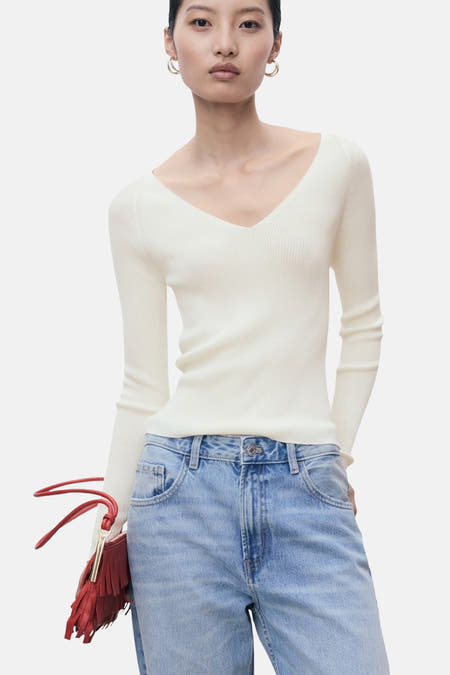 Pull blanc à col rond de MANGO, porté avec un pantalon de cuir bleu et une sacche rouge frangée.