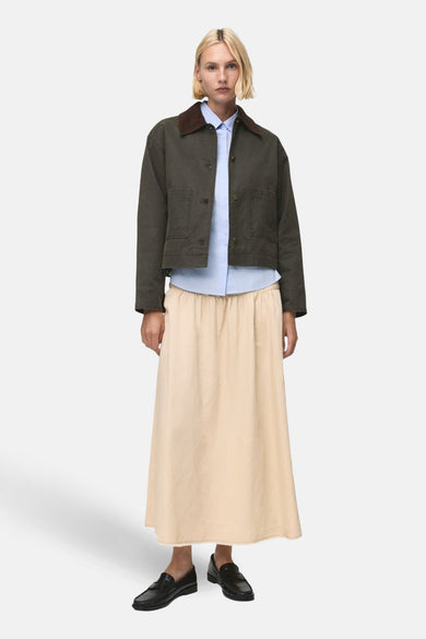 Veste kaki courte de MANGO, avec un col marron, portée avec une chemise bleu clair et une jupe beige.