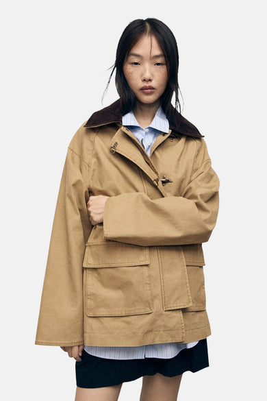 Manteau court beige de MANGO, avec un col marron et des poches à l'avant, porté sur une chemise rayée.