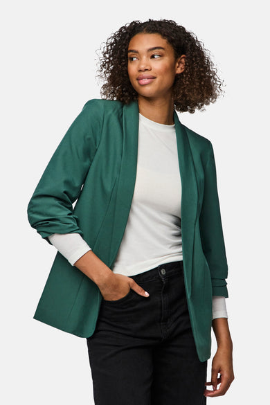 Groene blazer van PIECES, gedragen over een wit shirt en een zwarte jeans, met een hand in de zak.