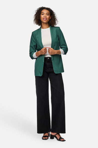 Blazer vert de PIECES, avec des manches longues et une ferverture ouverte, porté avec un chemise blanc, un pantalon noir et une poche main.