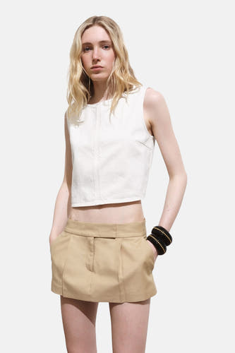 Blouse écru sans manches de MANGO, portée avec un mini-jupe kaki et des bracelets noirs avec des détails dorés.