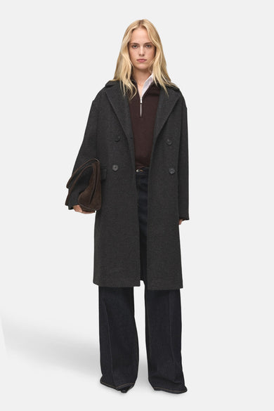 Manteau long gris de MANGO, avec une collier et et un fermeture à glissière argentée, porté sur une chemise blanche.