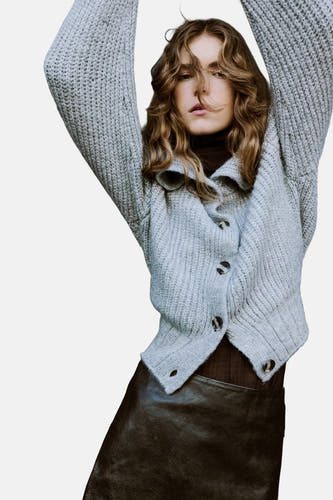 Gilet gris de MANGO porté sur une jupe en cuir, avec un sac marron et des bottes hautes.