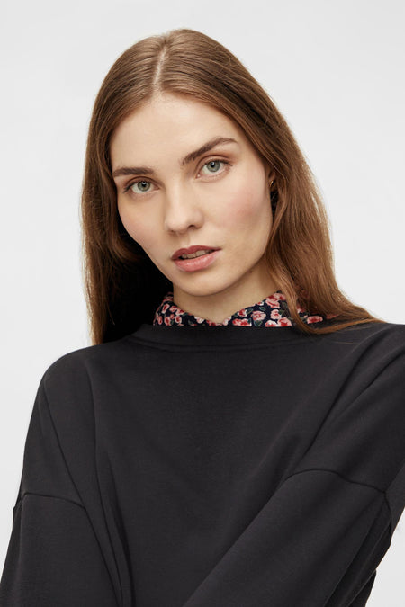 Gilet noir de PIECES, avec un col rond et des manches longues, porté sur une blouse à imprimé fleuri.