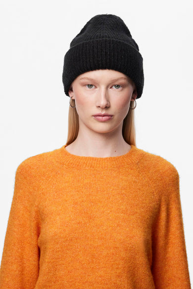 Casquette noire de PIECES®, à texture tricotée, portée avec des boucles d'oreilles en argent et un pull orange.