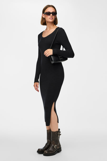 Robe noire à longues manches de PIECES®, avec une fente et portée avec des bottes brunes et un sac à main noir.