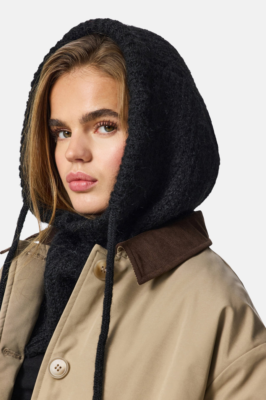 Chapeau noir de PIECES, avec une texture tricotée et des lanières, porté avec un manteau beige à col brun.