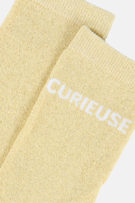 Gele gebreide sokken van PIECES® met glinstering en witte 'CURIEUSE' opdruk, in een diagonale close-up.