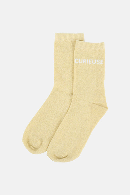 Chaussettes tricotées jaunes de PIECES avec paillettes, le texte 'CURIEUSE' et une étiquette blanche.