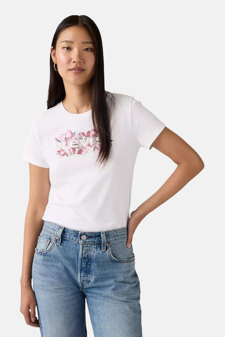 Vrouw in een witte T-shirt met bloemenprint en blauwe jeans, staand tegen een witte achtergrond.
