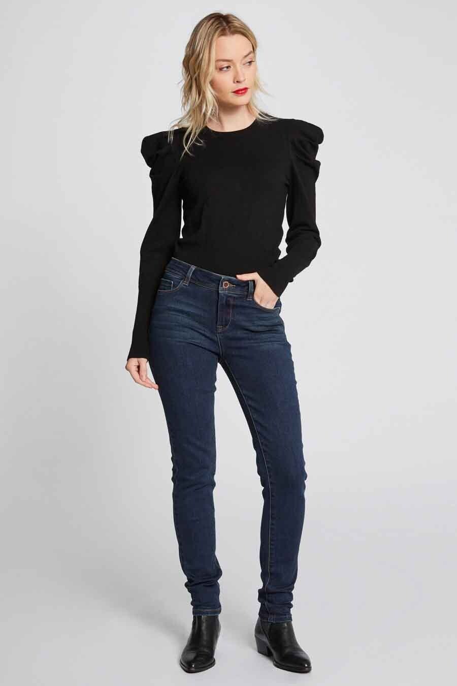Femme portant un pull noir et des jeans foncés avec des chaussures noires, debout.