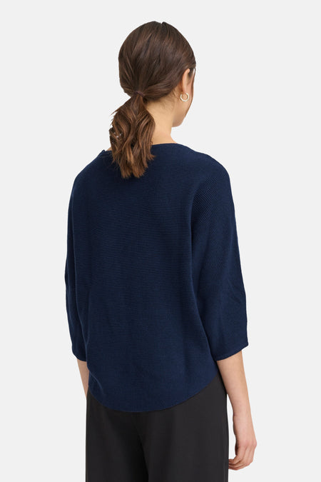 Blauwe gebreide trui, achteraanzicht, zwarte broek.
