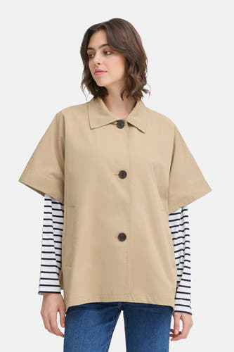 Manteau beige de France, avec des manches courtes, porté sur une chemise rayée et un jeans bleu.