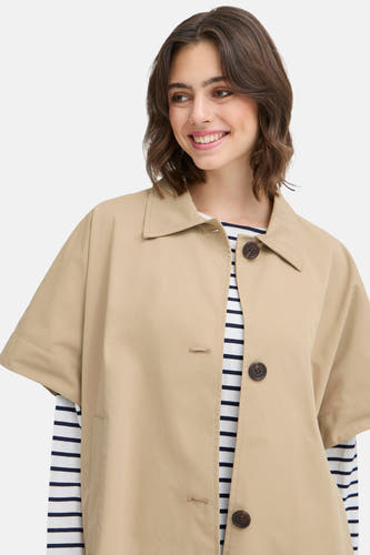 Manteau beige de France, avec des boutons et des manches courtes, porté sur un chemisier rayé.