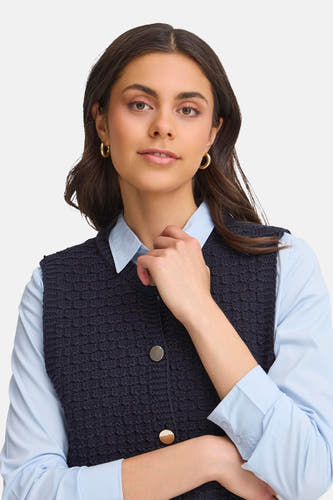 Gilet bleu de France, avec une fermeture à boutons sur un chemisier bleu clair, porté avec des boucles d'oreilles dorées.