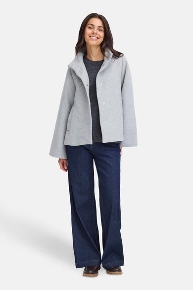 Manteau court gris de France, avec des manches longues et une coupe décontractée, associé à des jeans foncés et des chaussures marron.