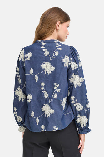 Blouse bleue à longues manches en Fransa, avec des fleurs blanches brodées et des manches bouffantes, portée avec une jupe noire.
