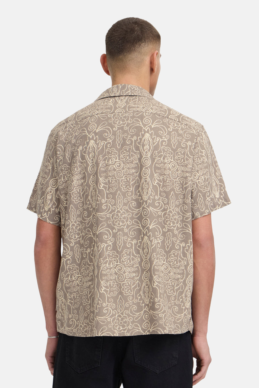 Chemise marron à manches courtes de SOLID, avec motif décoratif blanc cassé, portée par un homme vu de dos avec un pantalon noir et un bracelet argenté.