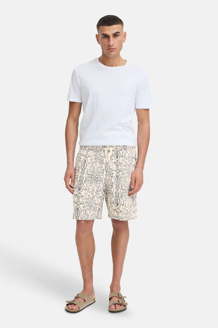 Crèmekleurige short met donkerblauw, paisley-achtig patroon van SOLID, met trekkoord. Gedragen met wit T-shirt en beige sandalen.