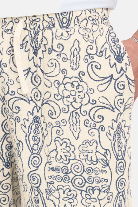 Short écru avec un motif bleu foncé de SOLID, avec taille élastique, cordon de serrage et une main avec un bracelet argenté dans la poche.