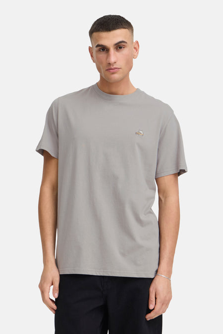 T-shirt à manches courtes taupe de SOLID avec un détail brodé sur la poitrine, porté avec un pantalon noir et des mocassins.