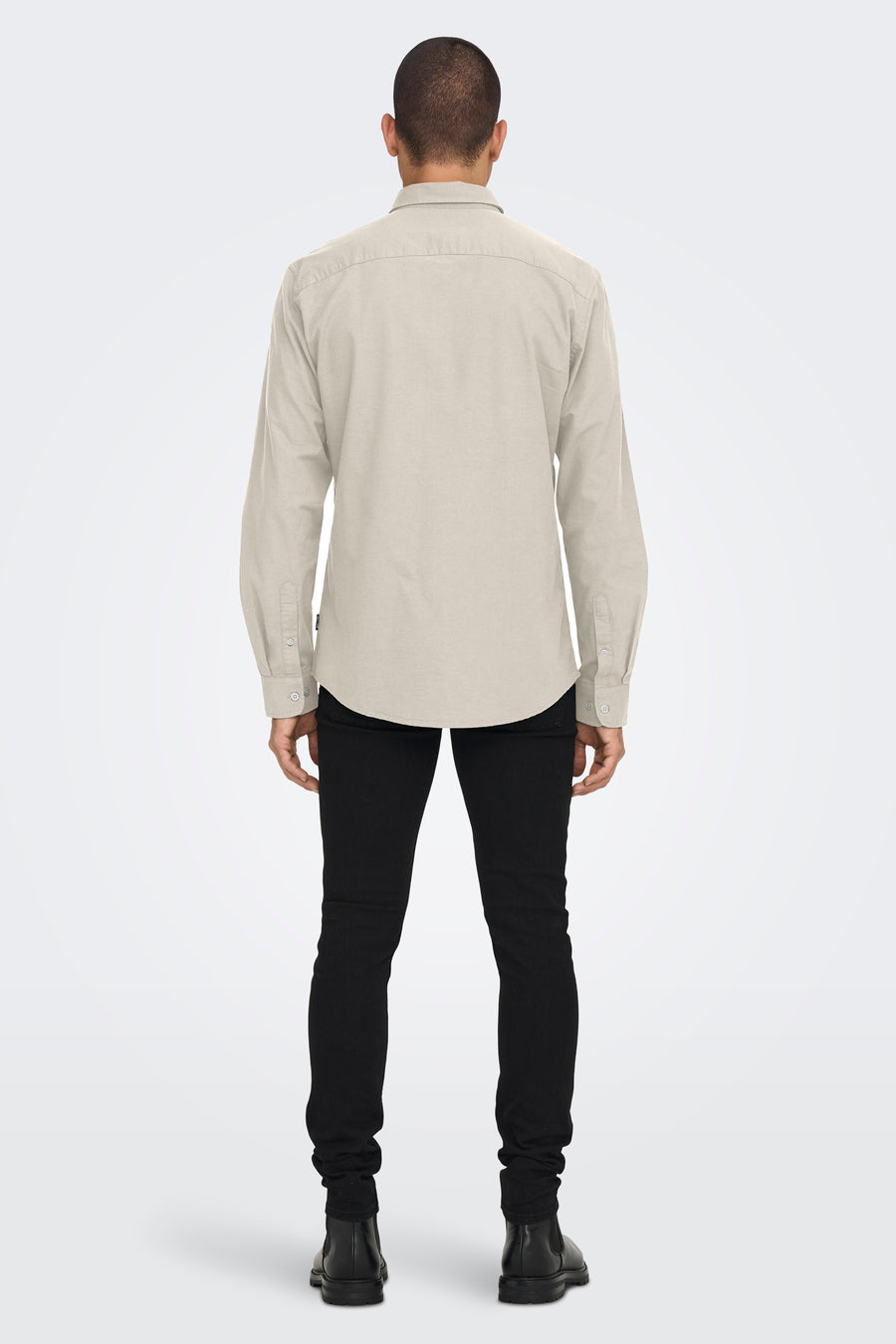 Achterkant van een man in een beige overhemd, zwarte broek en zwarte laarzen tegen een witte achtergrond.