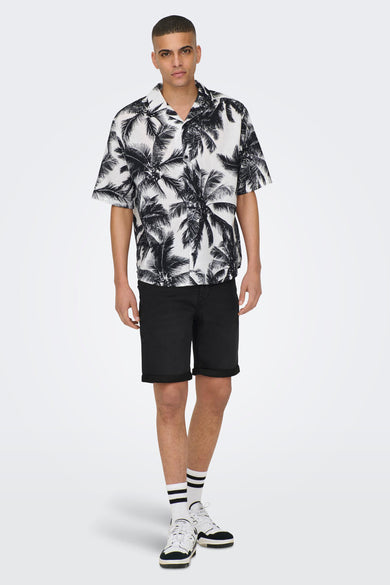 Homme en une chemise tropicale, un short noir, des chaussettes blanches et des baskets, debout sur un fond blanc.