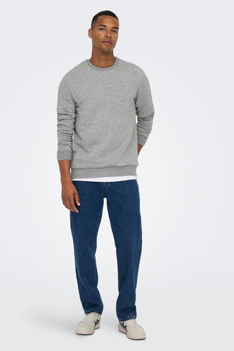 Homme en pull gris et en jeans bleus, debout contre un fond blanc.