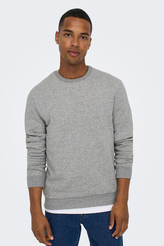 Un jeune homme pose dans un pull gris et des jeans bleus.