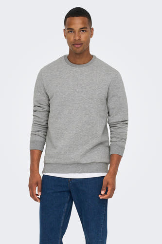 Homme en chemise grise et jeans bleus contre un fond blanc.