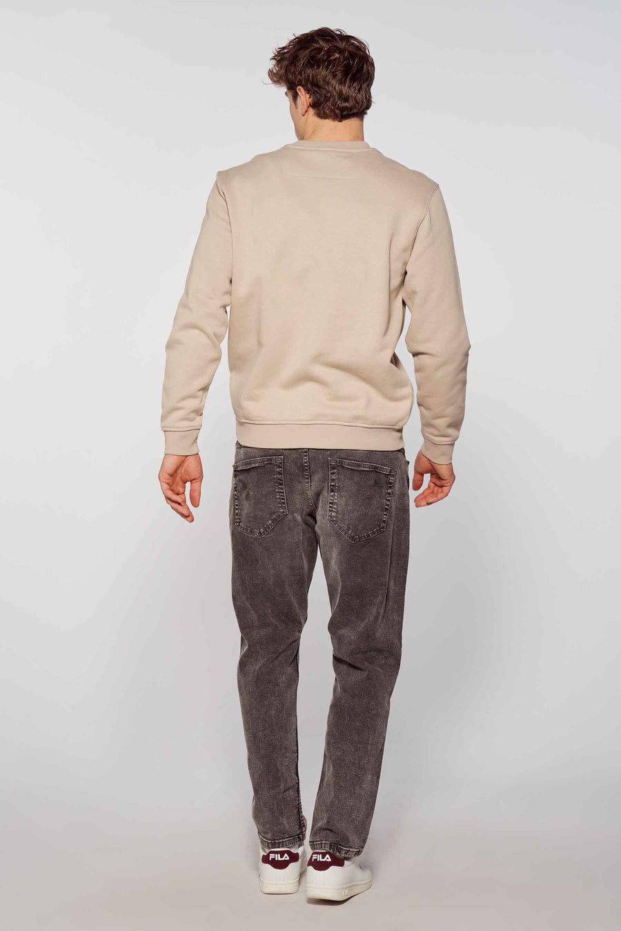 Homme assis, pull beige, jeans gris, baskets FILA blanches, arrière-plan clair.
