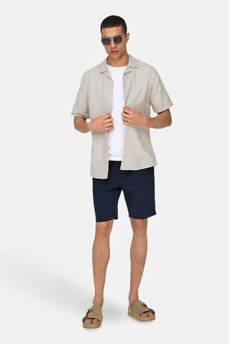 Chemise beige à manches courtes de ONLY & SONS, portée sur un T-shirt blanc, avec des shorts bleu marine et des sandales.