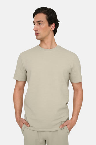 Man draagt beige T-shirt en broek, handen in zakken.
