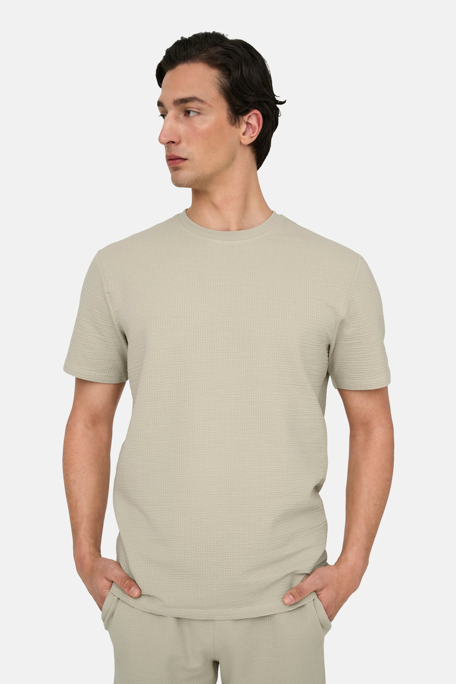 Man draagt beige T-shirt en broek, handen in zakken.