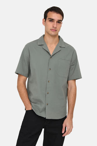 Chemise kaki à manches courtes de ONLY & SONS®, avec col boutonné et poche poitrine, portée avec un pantalon noir.
