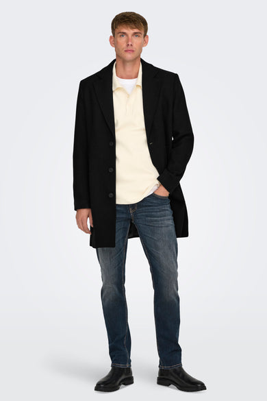 Long manteau noir d'ONLY & SONS porté sur une polo crème et des jeans bleu marine.