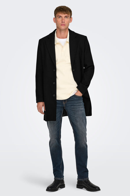 Long manteau noir d'ONLY & SONS porté sur une polo crème et des jeans bleu marine.