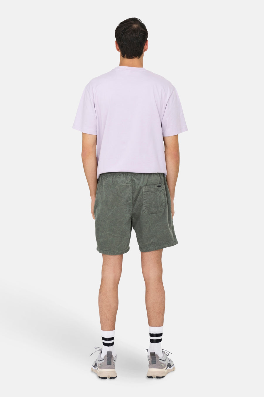 Man van achter, paars shirt, groene corduroy shorts, witte sokken met zwarte strepen, grijze sneakers.